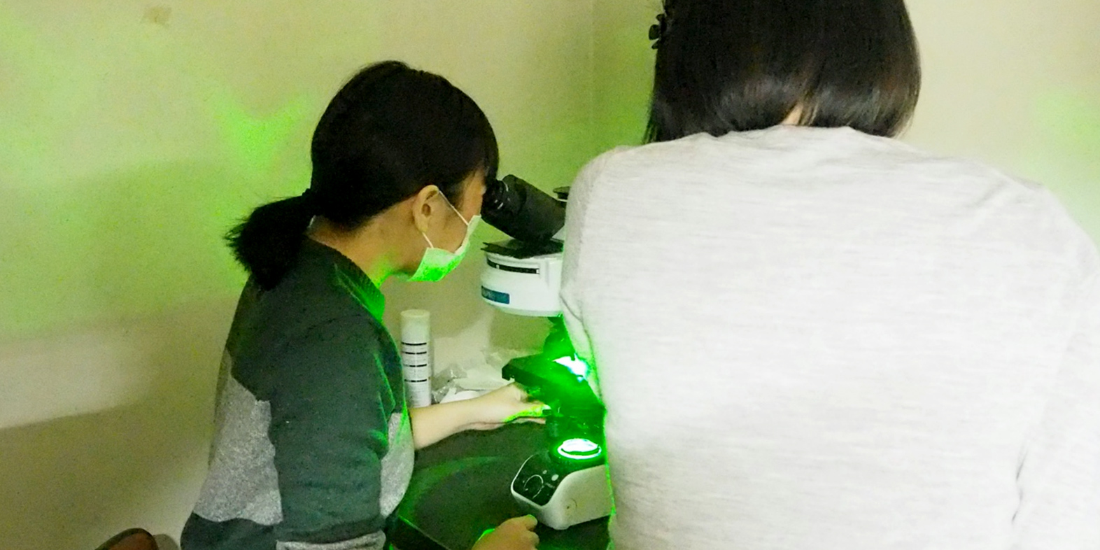 Ms. YAMAGUCHI peers into a fluorescence microscope as SA Assist. Prof. SUYAMA explains what she is seeing